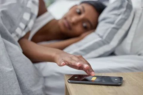 Woman in bed reaching to turn off alarm on smartphone on bedside table early morning