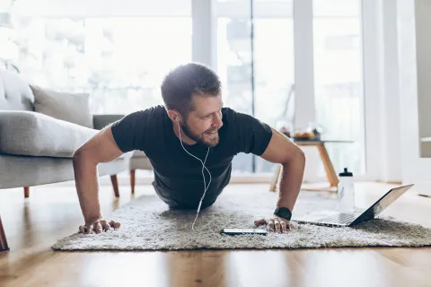 Man doing push-ups at home with earphones, laptop, and water bottle on wooden floor in bright living room.