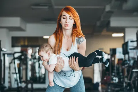 Young woman with red hair exercising by lifting a baby in a gym setting with workout equipment in the background