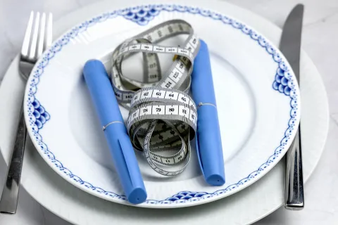 Blue insulin pens and a white measuring tape on a white plate with blue patterns, fork and knife beside.