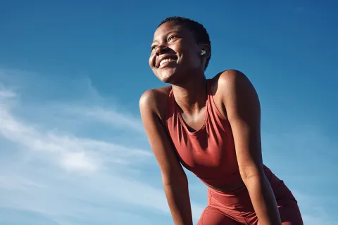 Smiling woman in red workout clothes wearing wireless earbuds outdoors under clear blue sky.