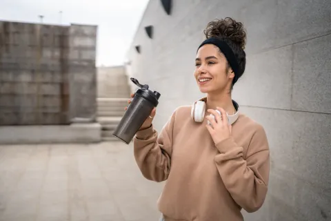 Smiling young woman with curly hair and headphones holds a black shaker bottle outdoors near concrete walls.