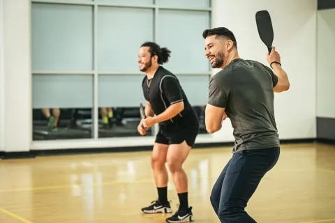 Two men playing pickleball indoors wearing athletic clothing and holding paddles on a wooden court.