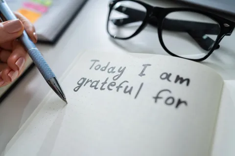 Person writing gratefulness journal entry with glasses and notebook nearby on white desk.