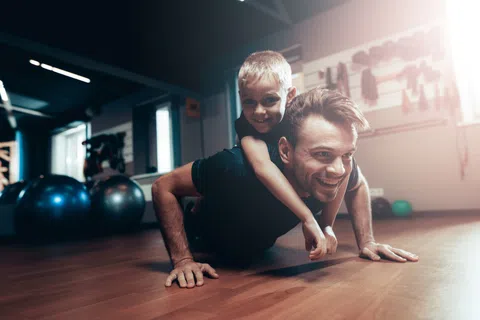Man doing push-ups with a young boy on his back in a bright gym setting, both smiling and having fun.