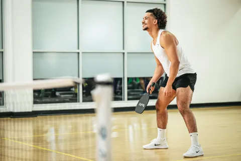 Athletic man in white tank top and black shorts playing pickleball indoors with paddle in hand.
