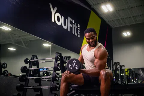 Smiling man lifting 40-pound dumbbell in YouFit gym with workout equipment and mirror in the background