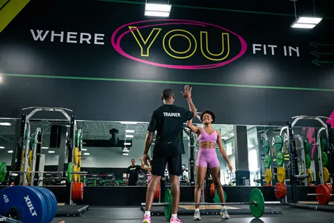 Gym trainer and woman in purple workout clothes giving high five by weightlifting area under motivational sign.