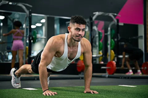 Man in white tank top doing push-ups on green turf in a gym with colorful weights and people in the background