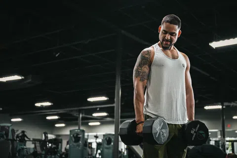 Man lifting dumbbells in YouFit Gyms wearing a white tank top and green pants under bright lights