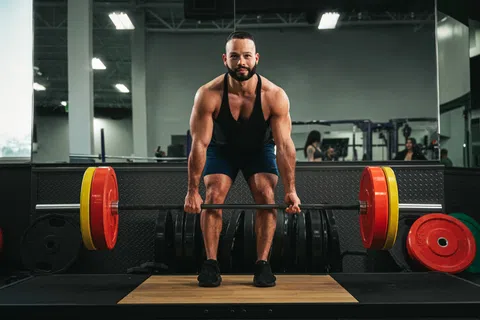 Muscular man wearing black tank top lifting heavy barbell with colorful weight plates in gym.