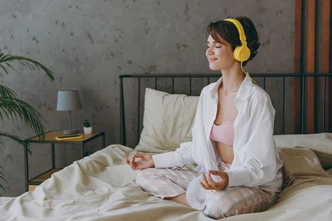 Young woman in pajamas meditating on bed with yellow headphones in cozy bedroom with soft lighting.