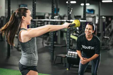 Woman lifting a kettlebell while a trainer encourages her in a gym setting with fitness equipment.