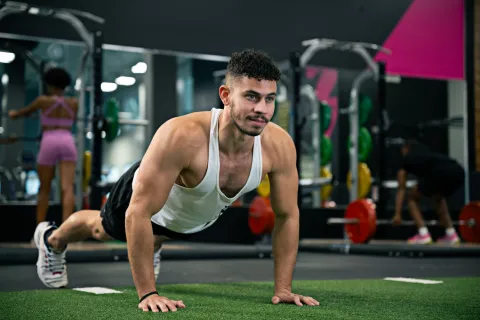 Man in white tank top doing push-ups on gym turf with weightlifting equipment and people in background