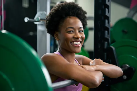Smiling young woman resting on a barbell in a gym with weight plates in the background, ready for weightlifting.
