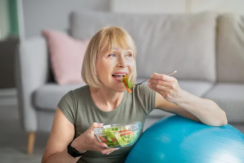 Older woman enjoying a healthy salad while sitting next to a blue exercise ball at home.
