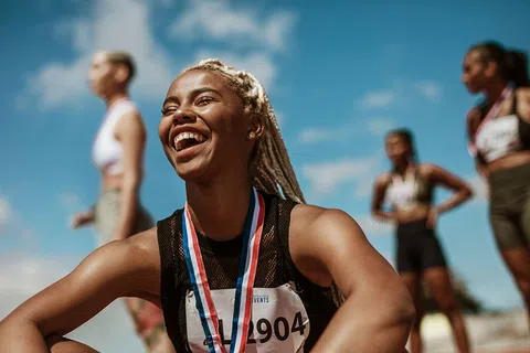 Smiling female athlete with medal around neck celebrating victory with fellow runners in background.