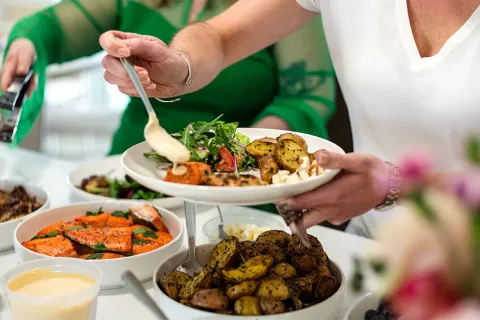 Person serving roasted potatoes and salad with grilled salmon at a buffet-style meal gathering.