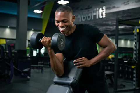 Smiling young man lifting a dumbbell using a preacher curl bench in a modern gym setting.