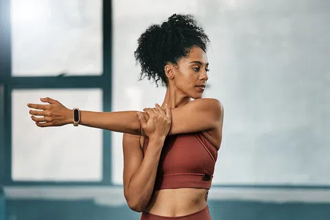 Woman in rust sportswear stretching her arm indoors with natural light from window.