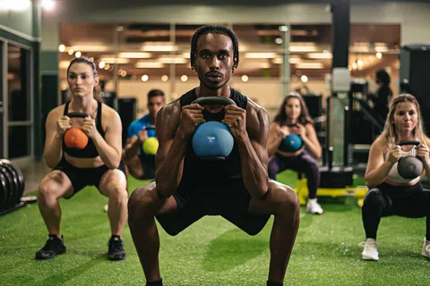 Group of diverse people doing kettlebell squats together in a gym with green turf flooring