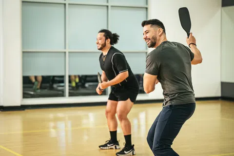Two men playing pickleball indoors with paddles on a polished wooden court.