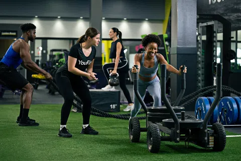 Group of diverse people exercising with battle ropes, sled push, and kettlebells in modern gym setting.