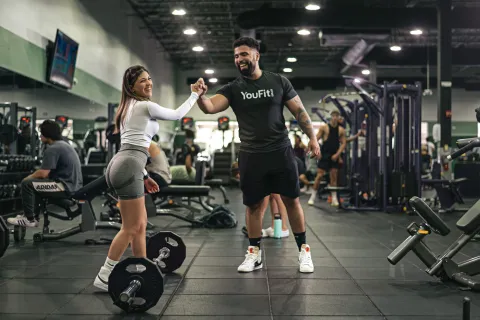 Two gym-goers fist bump in a busy fitness gym with weights and exercise equipment around.