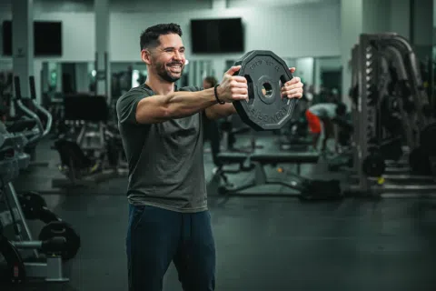 Smiling man lifting a weight plate in a modern gym with various exercise equipment in the background