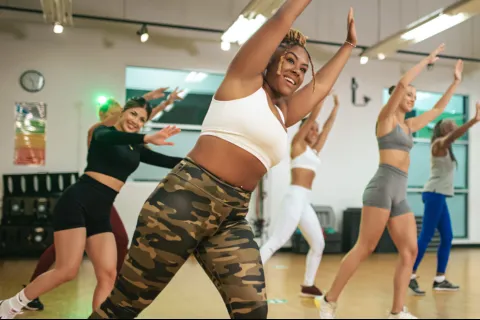 Diverse group of women in activewear smiling and stretching arms up during a fitness class in a studio.