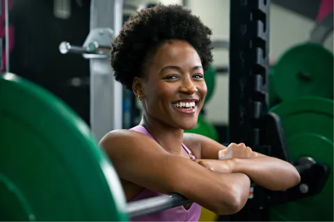 Smiling woman resting on a barbell rack in a gym surrounded by weight plates and fitness equipment.