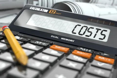 Close-up of a calculator displaying the word COSTS with rolled blueprints and a pencil nearby.