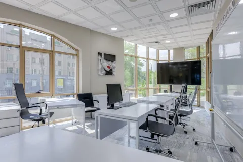 Modern bright office space with white desks, black chairs, large windows, computer monitors, and a wall-mounted TV.