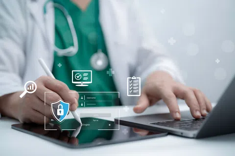 Medical professional using a digital tablet and laptop with cybersecurity and data icons overlayed on screen.