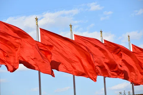 Multiple red flags waving on gold-tipped flagpoles against a blue sky with clouds.
