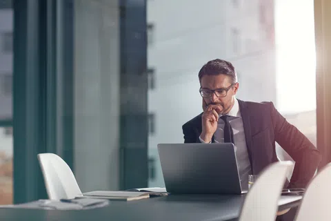 Businessman in suit working thoughtfully on laptop in modern office with large windows and natural light.