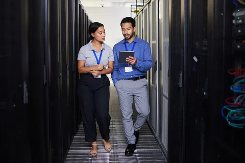 Two IT professionals walking and discussing data on a tablet between server racks in a data center.