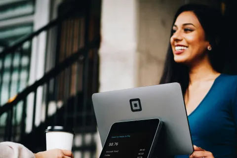 Smiling woman operating a Square point-of-sale system with a customer holding a coffee cup in a cafe.