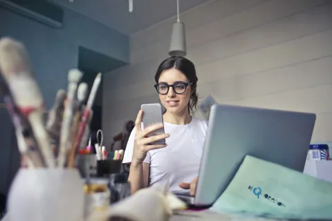 Young woman with glasses using smartphone and laptop in casual workspace with art supplies in foreground
