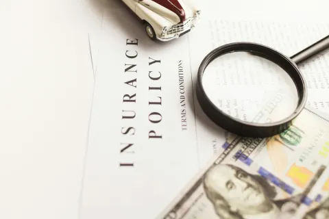 Magnifying glass on insurance policy with dollar bills and a vintage toy car on white surface.