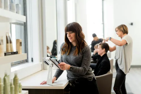 Woman checking tablet at salon counter while hairstylist cuts customer hair in modern salon setting