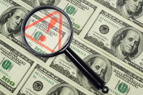 Magnifying glass highlighting red warning symbol on multiple hundred dollar bills beneath it.