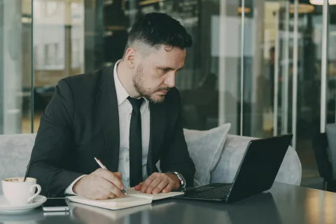 Businessman in suit working on laptop and writing notes at a cafe table with coffee cup.