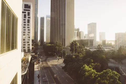 Early morning cityscape with tall buildings, empty streets, and lush green trees under soft sunlight.