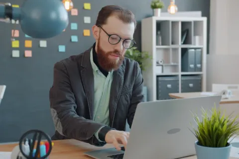 Man with glasses working on a laptop in a modern office with sticky notes and plants.