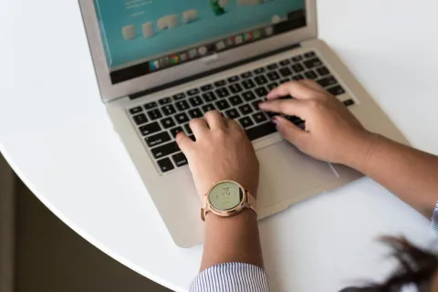 Person typing on a laptop with a smartwatch showing time 11:24 on their wrist over a white table.