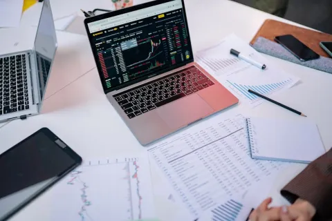 Laptop displaying stock market charts and data surrounded by financial reports, notebook, and smartphone on desk.