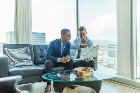 Two young businessmen in a modern office discussing work on a laptop with floor-to-ceiling windows and city view.