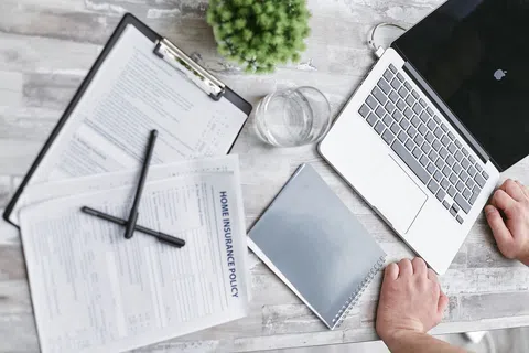 Workspace with laptop, home insurance policy documents, pens, notebook, glass of water, and small plant on wooden table.