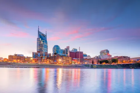 Nashville skyline at dusk with glowing city lights reflecting on calm river under a colorful sky.